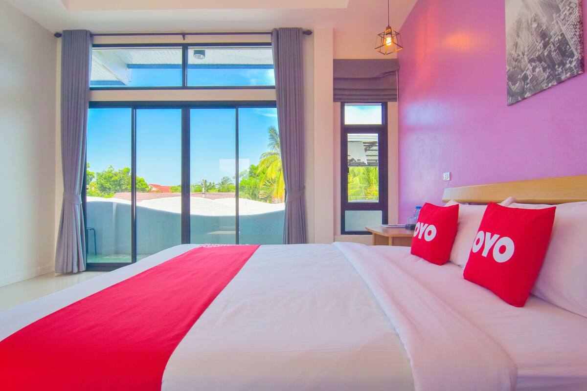 A hotel room with a large bed, two red pillows, a red throw, and a cityscape view through large windows.