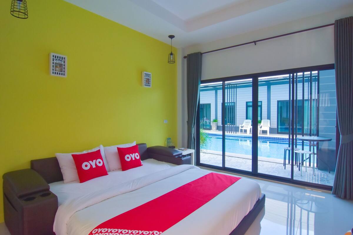 A hotel room with a large bed, a bright yellow wall, and a view of a swimming pool and outdoor seating area.