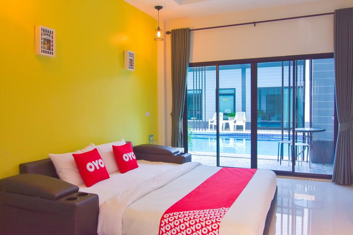 A hotel room with a large bed, a bright yellow wall, and a view of a pool and outdoor seating area.