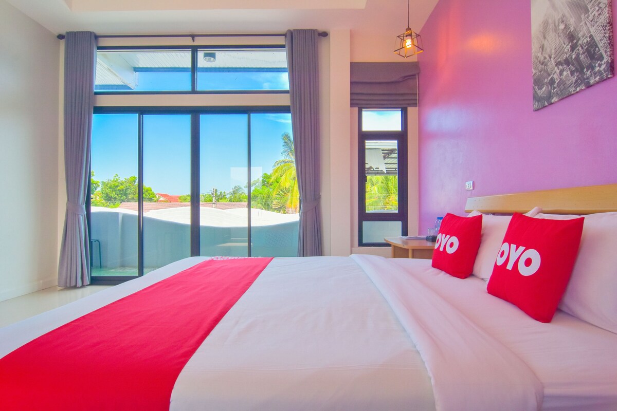 Habitación de hotel con una cama grande, dos almohadas rojas, una manta roja y vistas a la ciudad a través de ventanas amplias.