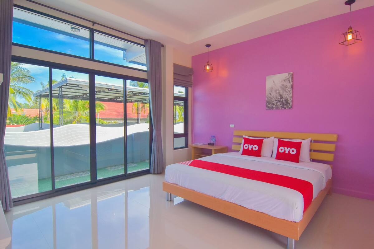 A modern hotel room with a large bed, a view of palm trees, and a bright pink wall.