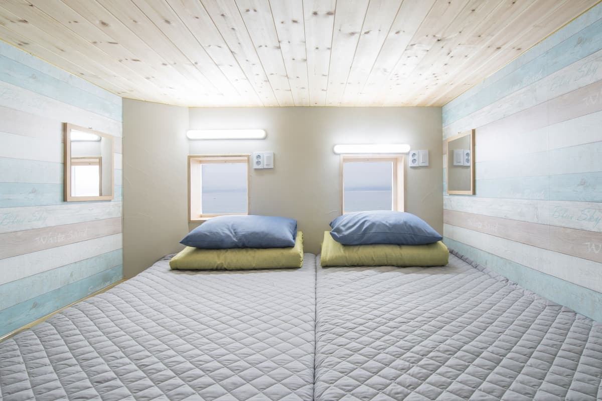 Ein Zimmer mit zwei Betten, jeweils mit einem Kopfkissen, einem Fenster und einem Spiegel an der Wand.