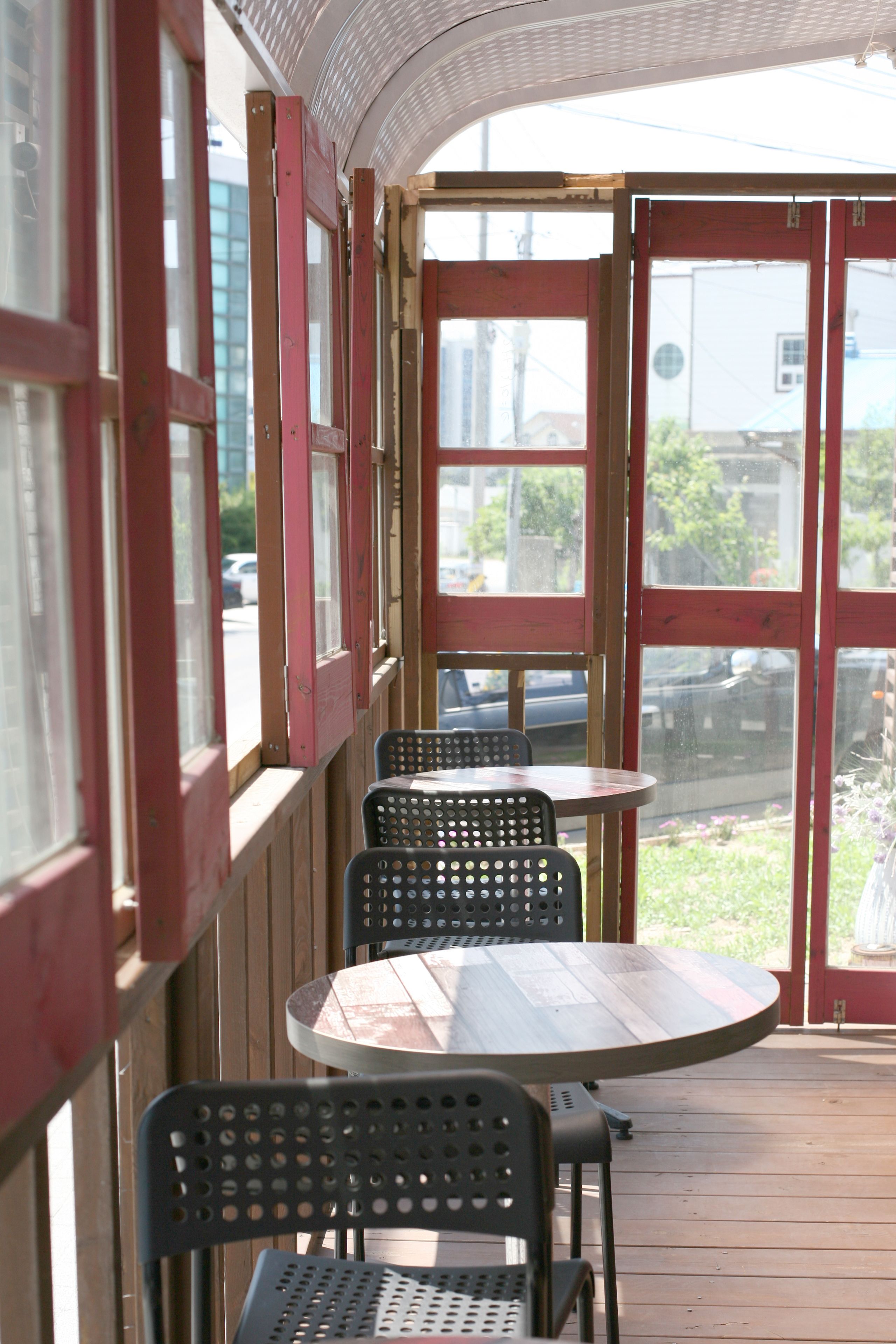 A covered outdoor seating area with wooden tables and metal chairs, large windows, and a view of the street.