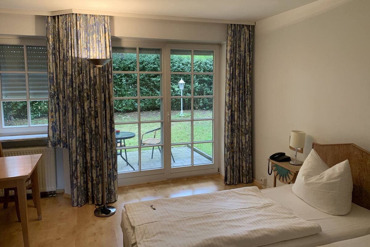 A hotel room with a bed, a desk, and a view of a garden through a glass door.