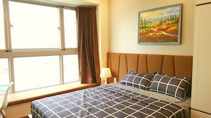 A bedroom with a large window, a bed with a checkered bedspread, a nightstand with a lamp, and a painting on the wall.