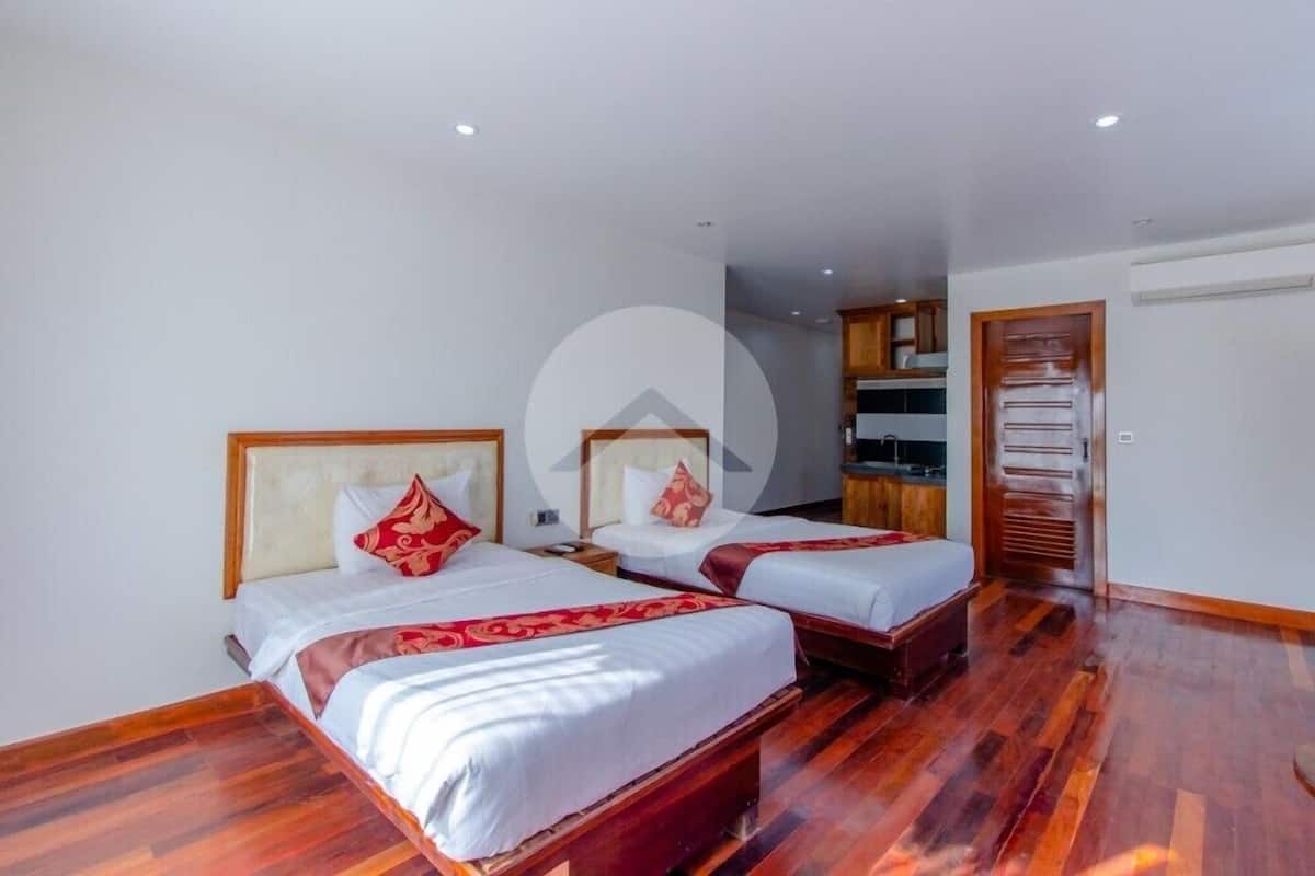 A hotel room with two beds, wooden flooring, a kitchenette, and a bathroom with a shower.