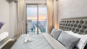 Une chambre moderne avec un grand lit, une tête de lit capitonnée et une vue sur la ville, visible à travers une porte coulissante.