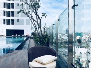 Uma área de piscina no terraço com vista para a cidade, uma cadeira de vime com almofadas e um guarda-corpo de vidro.