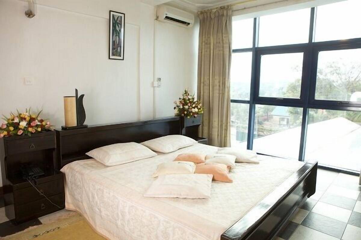 A neatly made bed with pillows, a bedside table with a vase of flowers, and a large window with curtains.