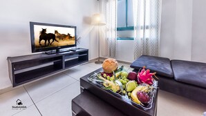 A living room with a TV displaying a horse race, a coffee table with fruits and a bread roll, a sofa, and a window with curtains.