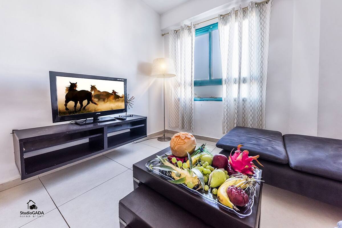 Una sala de estar con un televisor que muestra una carrera de caballos, una mesa de centro con frutas y un pan, un sofá y una ventana con cortinas.