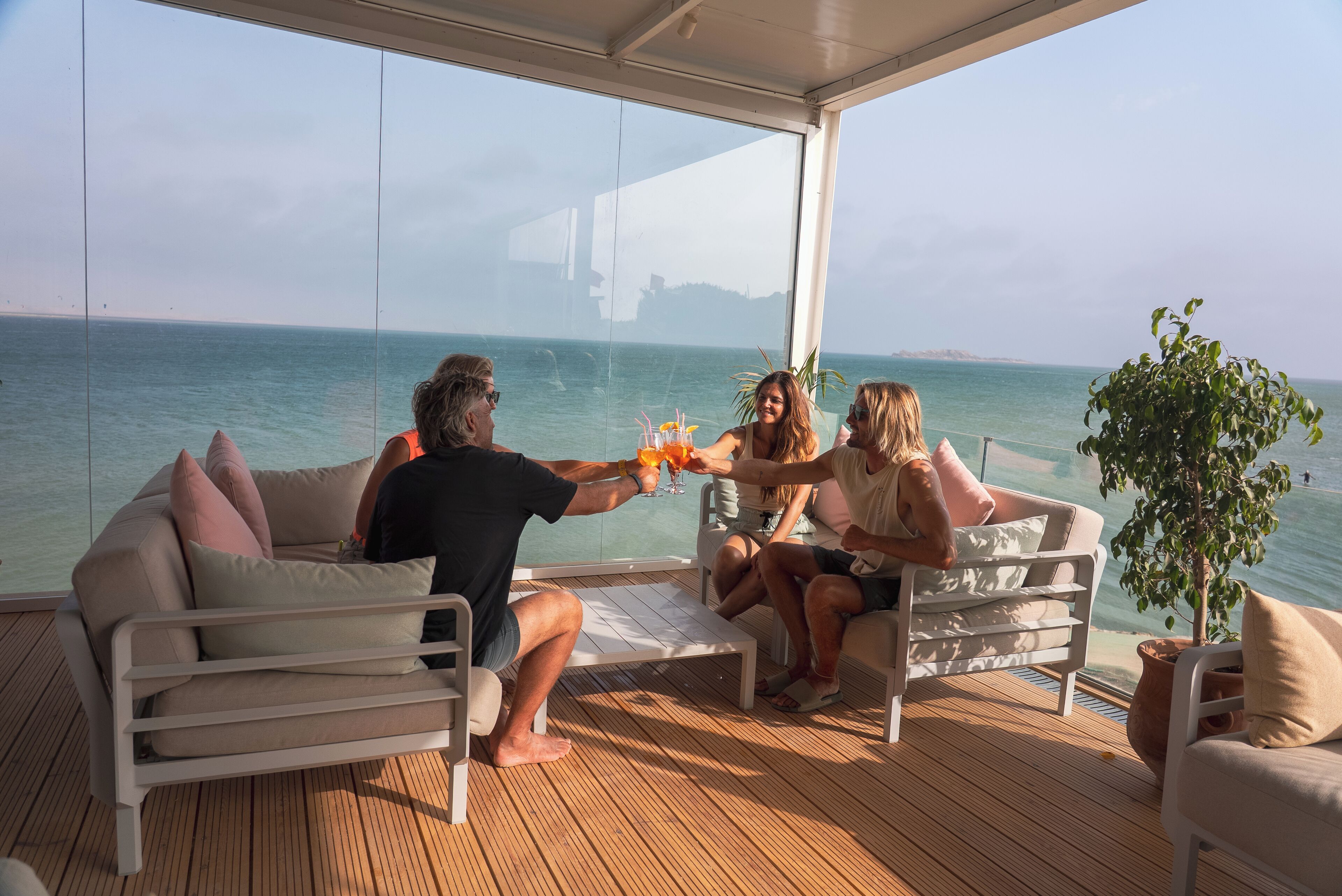 Een groep mensen proost met drankjes op een dek met uitzicht op zee.