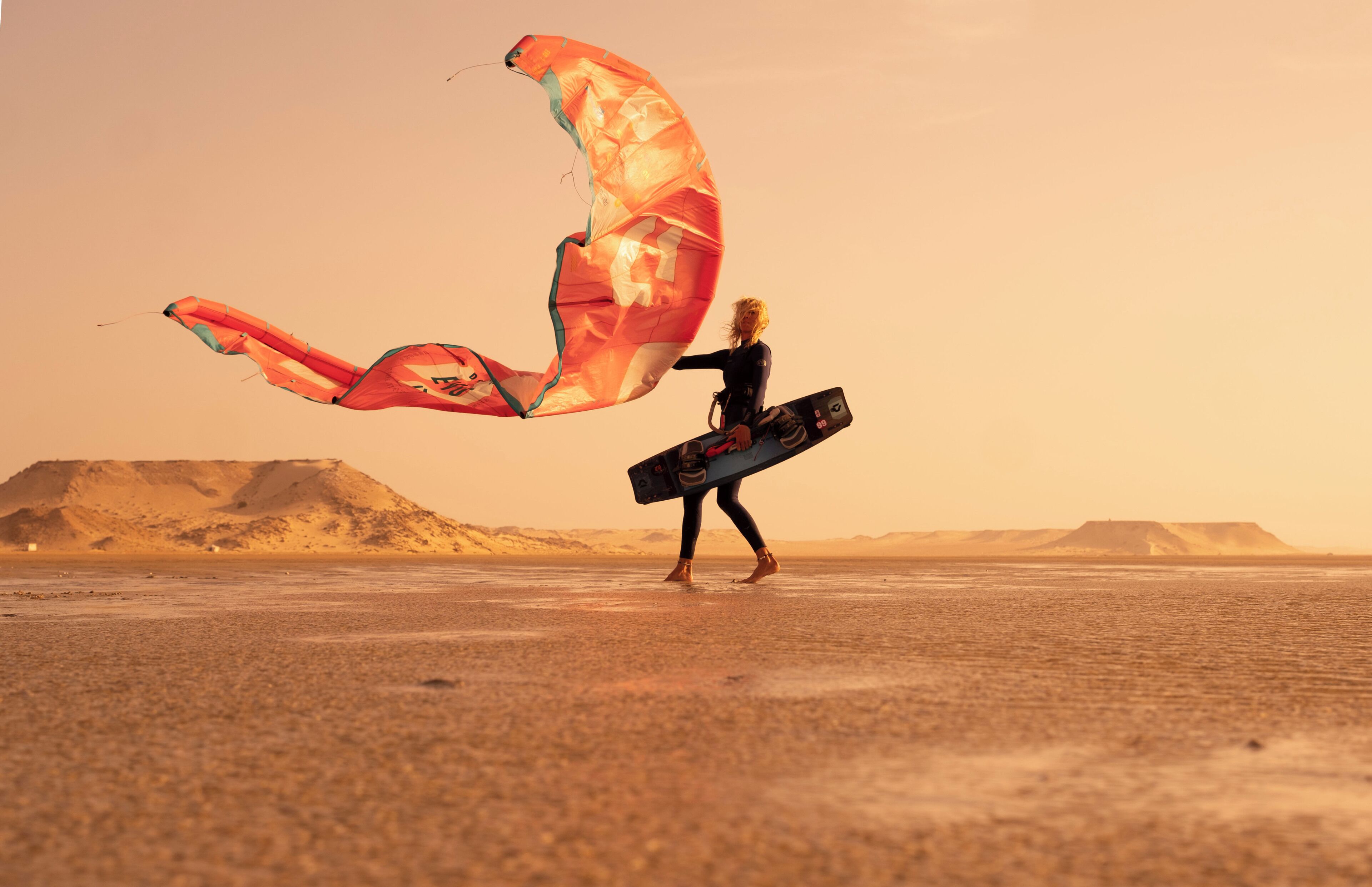 A person with a large kite and a surfboard on a barren landscape.