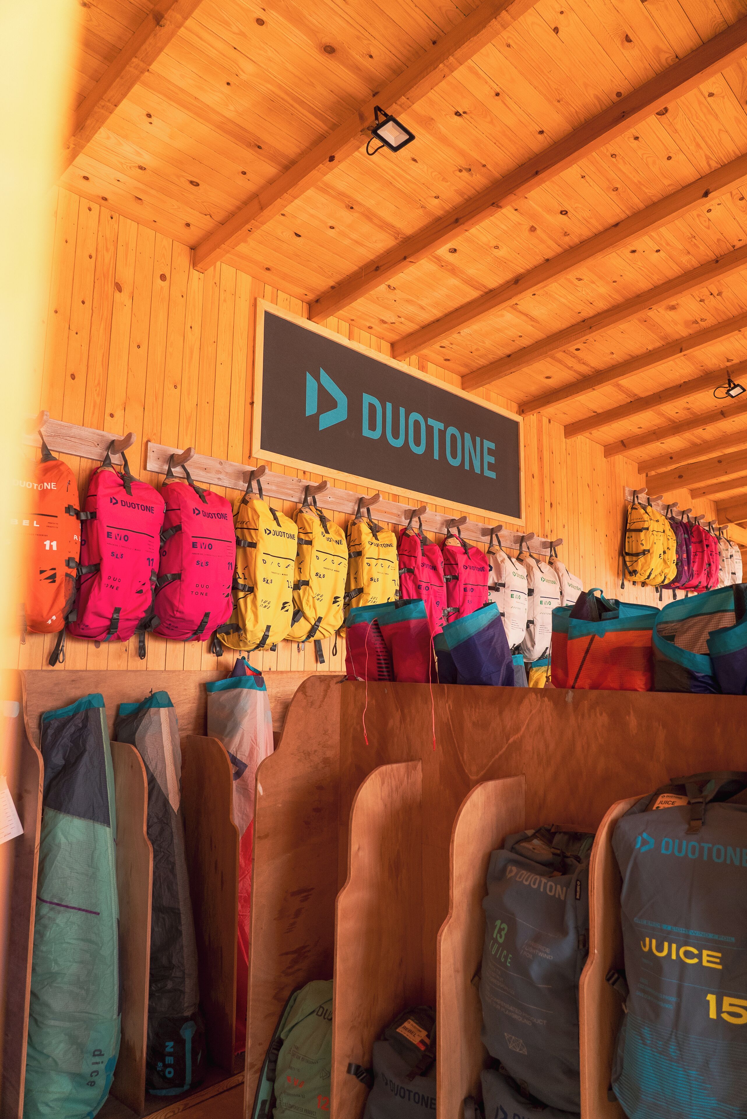 A room with wooden walls and ceiling, featuring a sign that reads "DUOTONE". There are multiple life jackets hanging on the wall and several bags with "DUOTONE" branding on the floor.