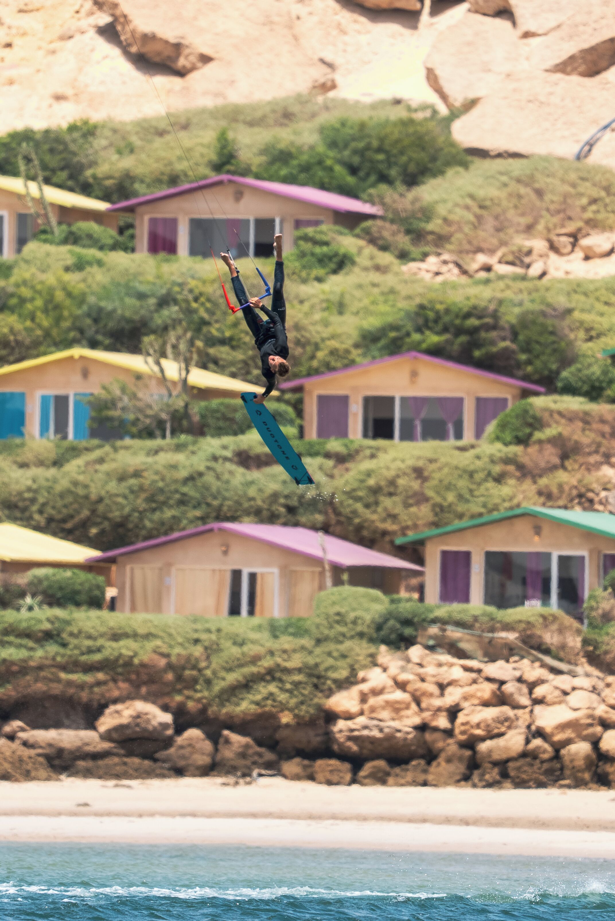Een persoon voert een truc uit op een surfplank, met een strand en huizen op de achtergrond.