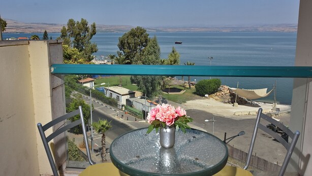 A balcony with a table and chairs, a vase with flowers, and a view of a body of water and distant landscape. - Family Vacation by Kinneret All Comforts (Tiberias)