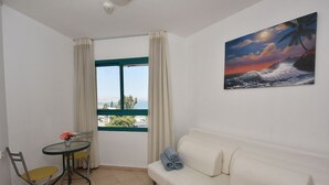 A room with a white sofa, a small round table with a flower vase, two chairs, a window with curtains, and a painting of a sunset over the ocean. - Family Vacation by Kinneret All Comforts (Tiberias)