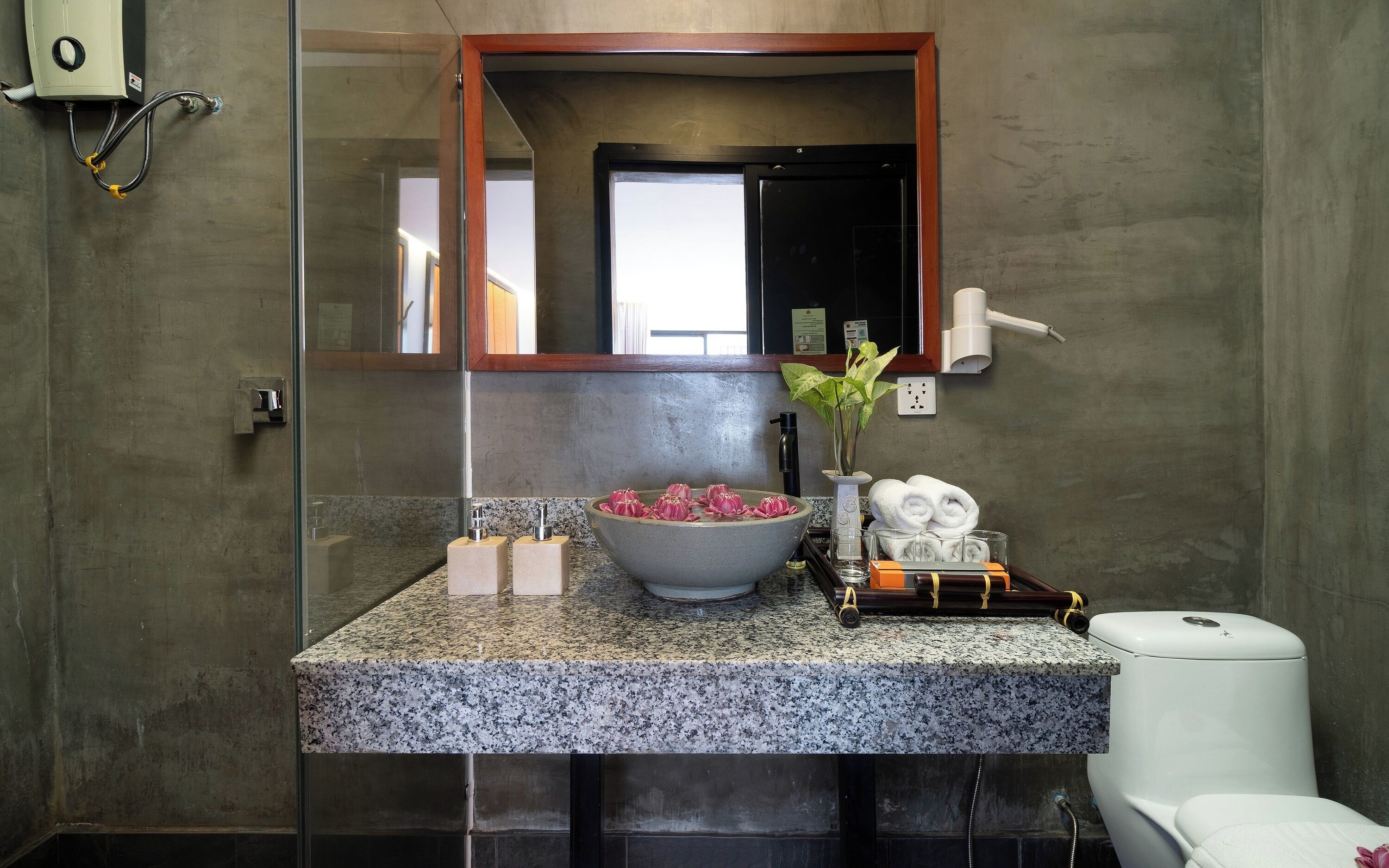 A bathroom with a granite countertop, a large bowl with pink flowers, a white toilet, and a mirror above the sink.