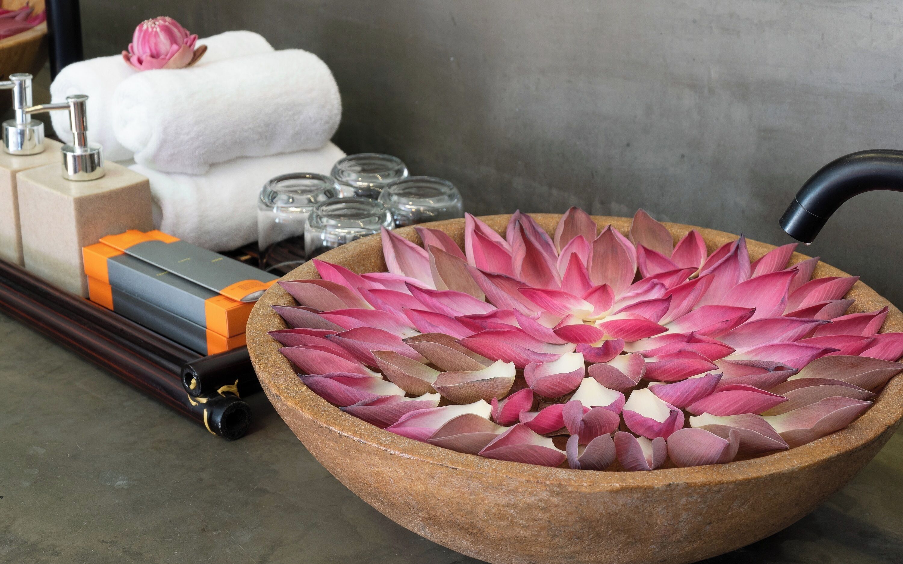 Un grand saladier en bois rempli de fleurs de lotus roses, une serviette blanche et un robinet noir.