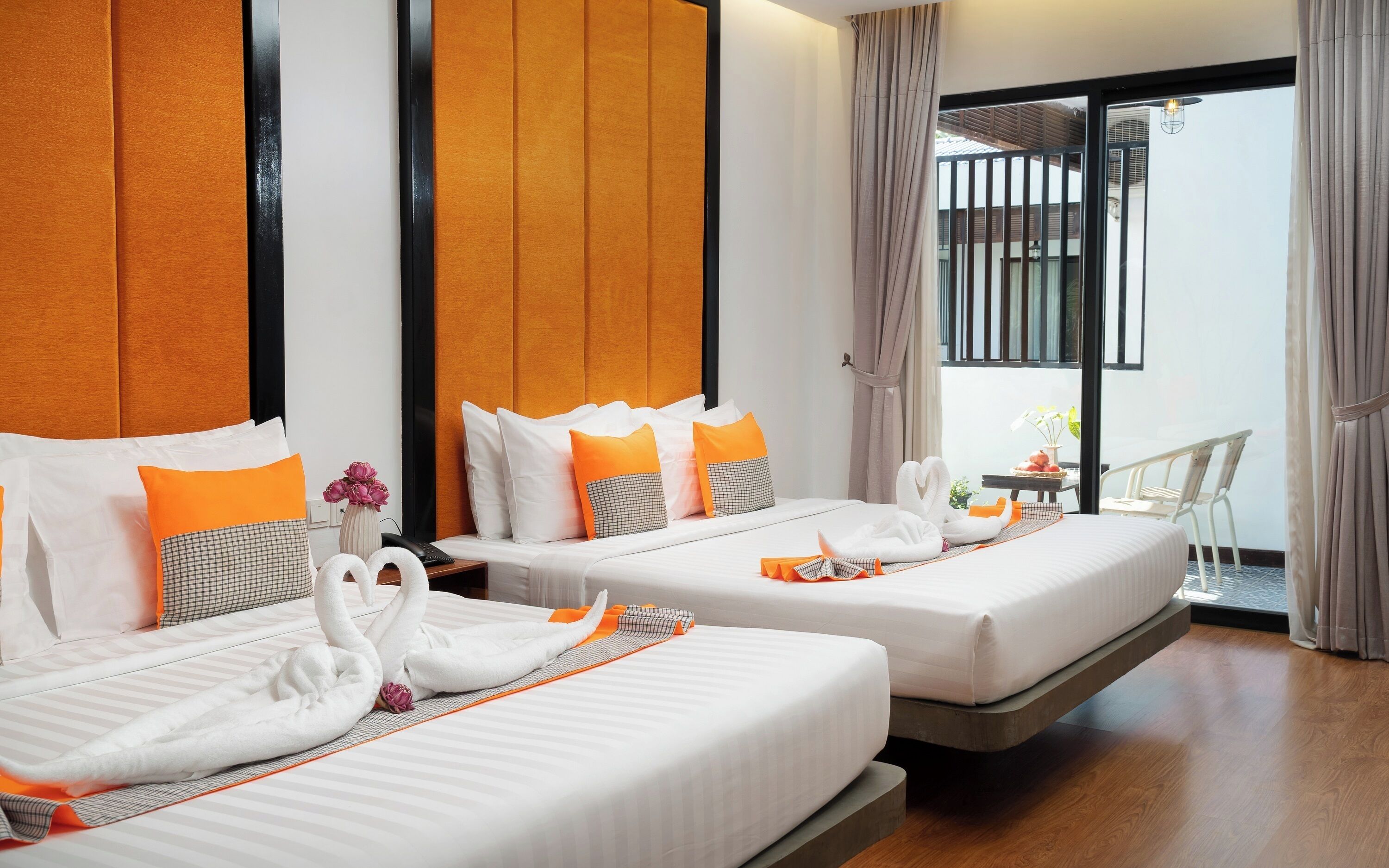 Habitación de hotel con dos camas, cada una con ropa de cama blanca y detalles en naranja, una mesita con un florero y vistas a un balcón con barandilla.