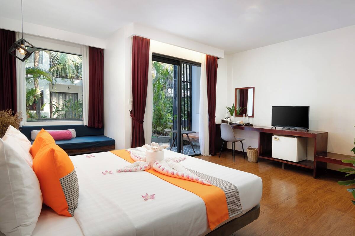 A hotel room with a bed, a desk, a television, and a view of greenery outside.