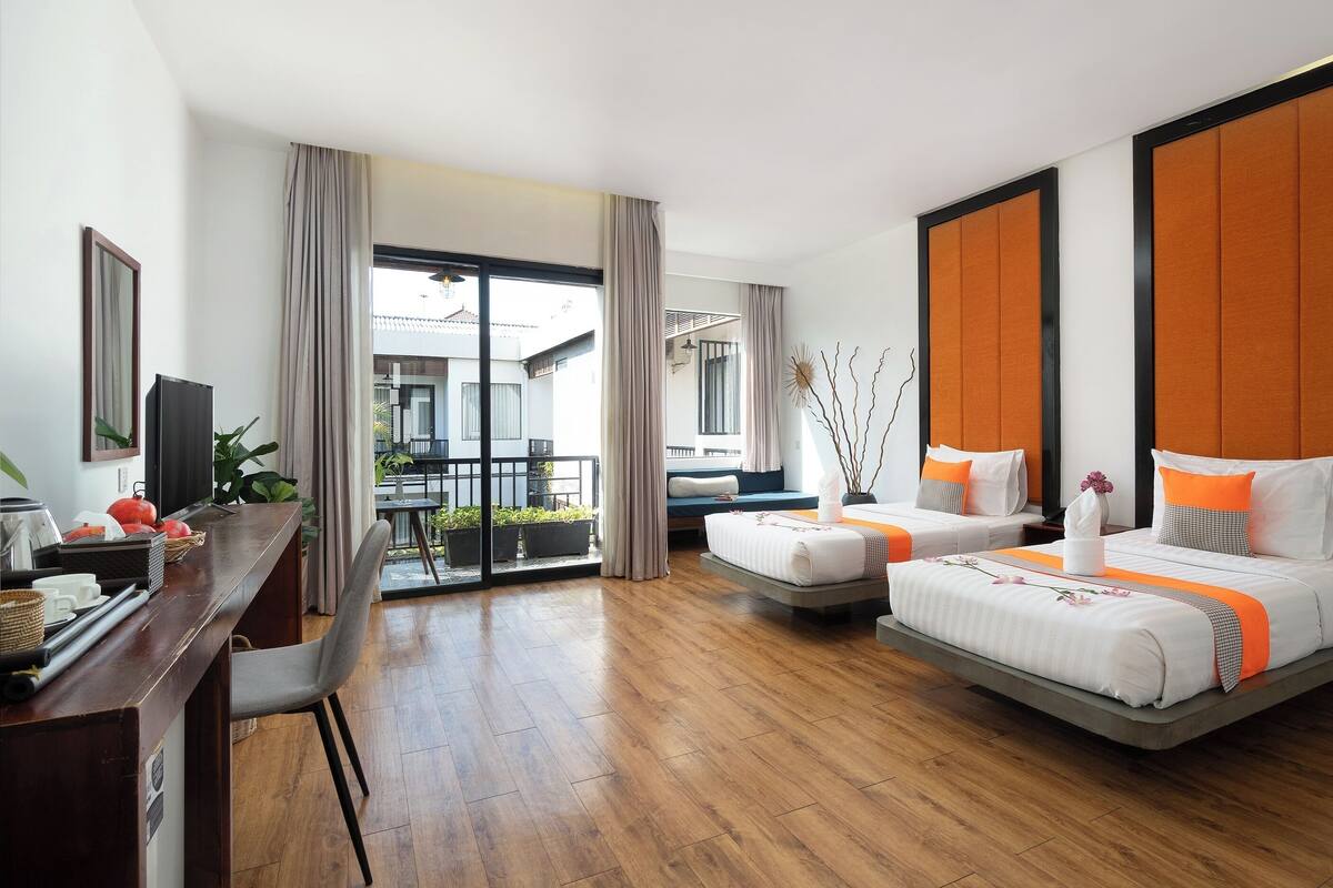 A hotel room with two beds, a desk, a TV, and a balcony with plants.