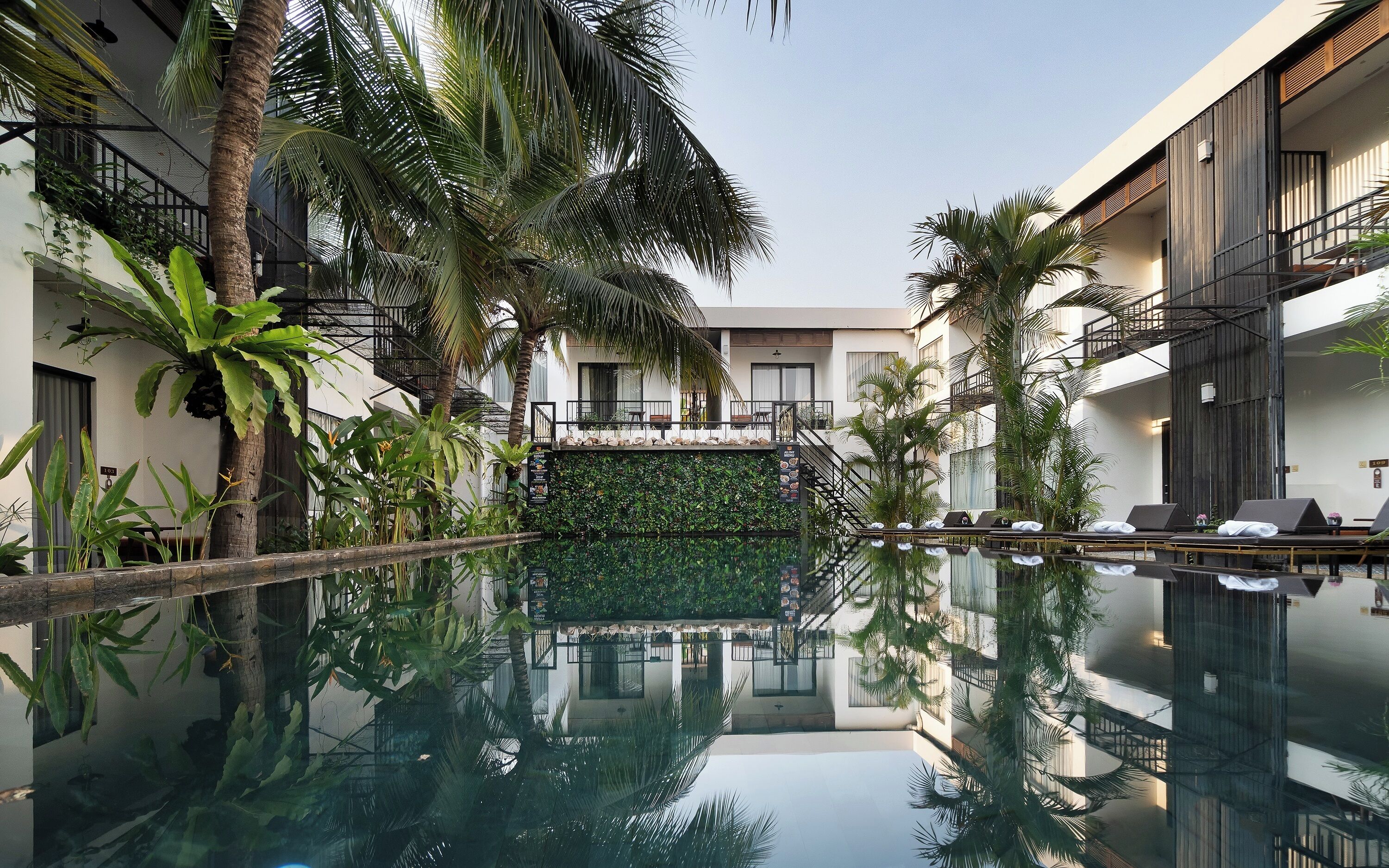 A modern hotel with a central pool, surrounded by lush greenery and palm trees.