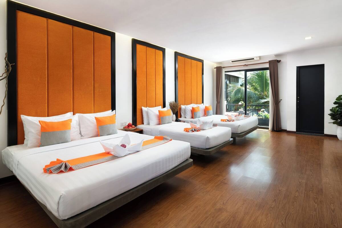 A hotel room with three beds, each with white bedding and orange accents, large windows, and a view of greenery outside.