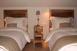 Quarto de hotel com duas camas, cada uma com cabeceira e almofada com a palavra "REVE". Uma pequena mesa de cabeceira de madeira com uma luminária fica entre as camas.