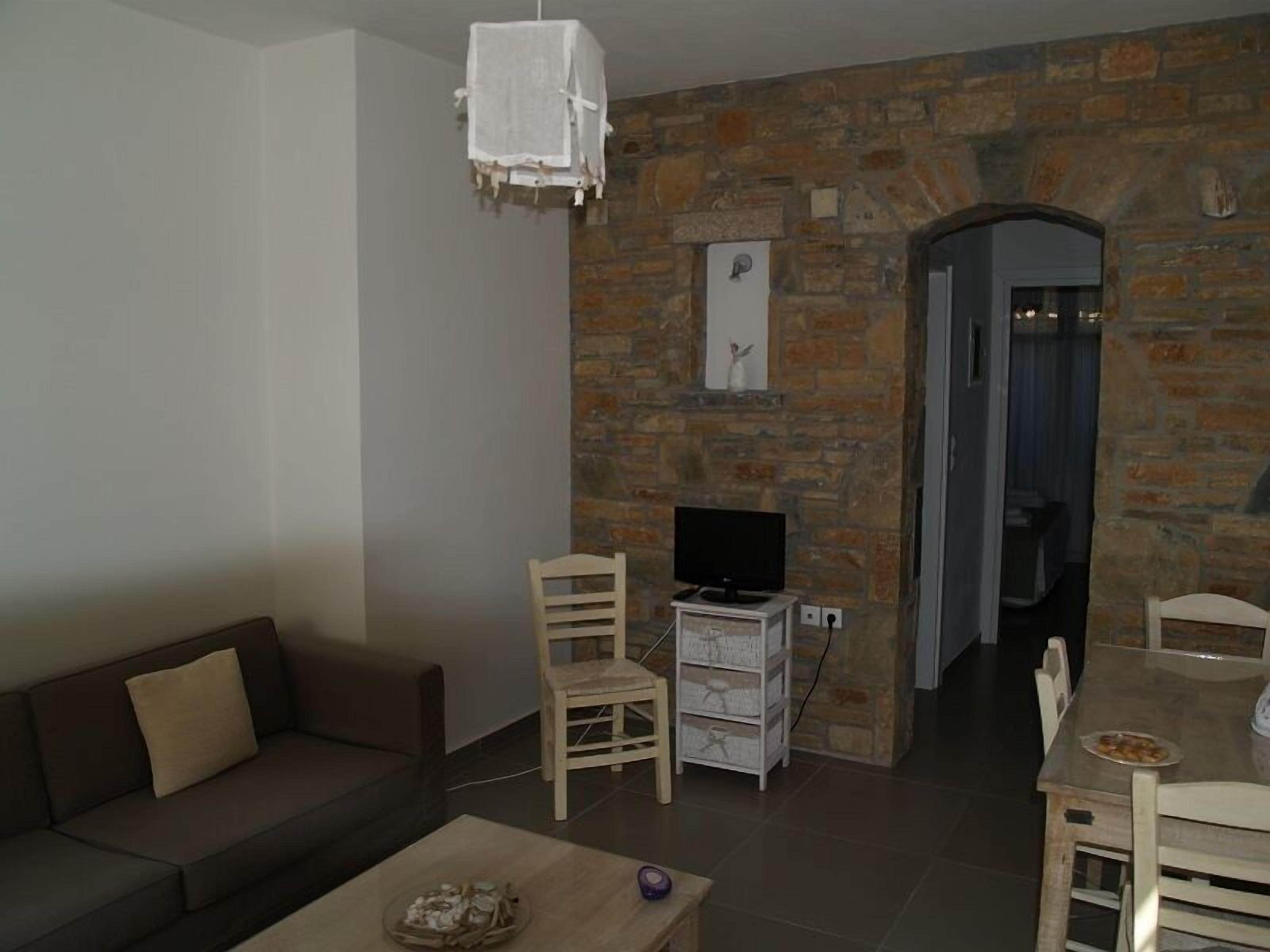 Una sala de estar con pared de piedra, un televisor sobre un mueble bajo, un sofá, una mesa de centro y un comedor con una mesa y sillas.