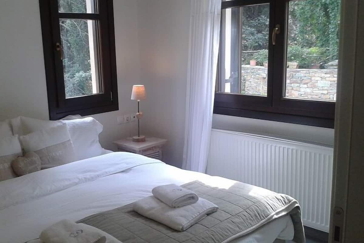 Una cama bien tendida con almohadas y una toalla doblada, una lámpara de noche y una ventana con vistas al verdor.