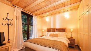 Un dormitorio acogedor con techo de madera, una cama grande con colcha marrón, lámparas de noche y una ventana con cortinas.
