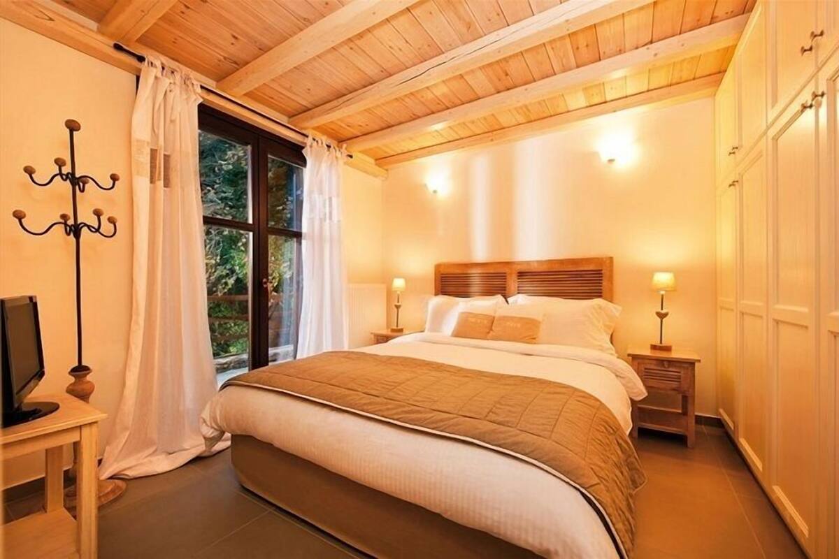Un dormitorio acogedor con techo de madera, una cama grande con colcha marrón, lámparas de noche y una ventana con cortinas.