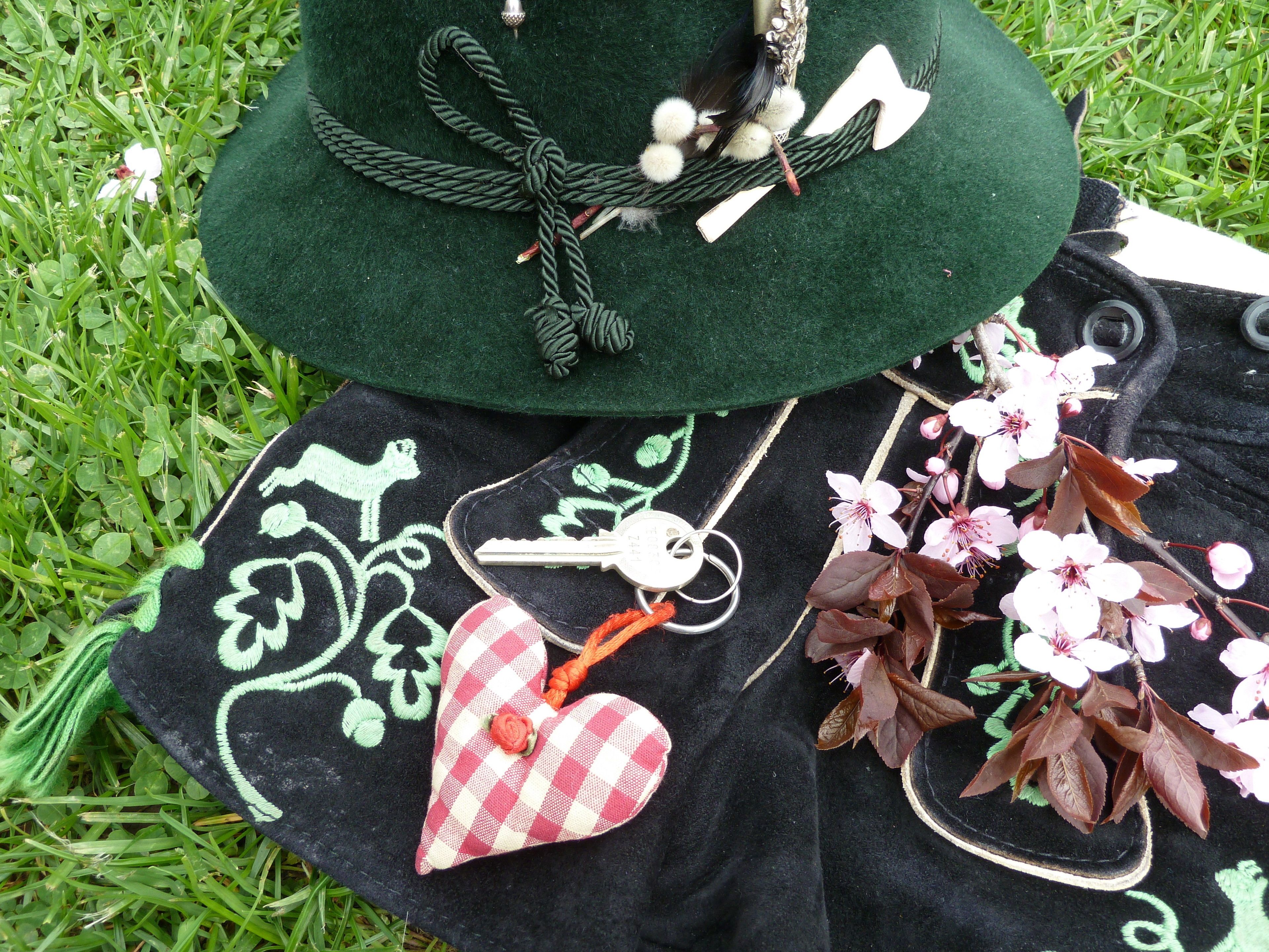 Un chapeau vert en feutre orné d’un ruban et d’une plume blanche, un tissu à carreaux noirs et blancs avec un motif en forme de cœur, et des fleurs de cerisier roses sur un tissu vert.