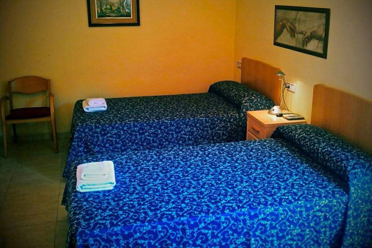 A room with two beds, a chair, a nightstand with a lamp, and two framed pictures on the walls.
