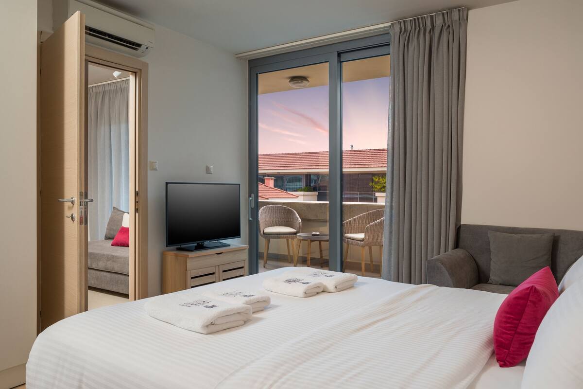 Habitación de hotel con una cama, televisión, balcón con sillas y puerta corrediza de vidrio.