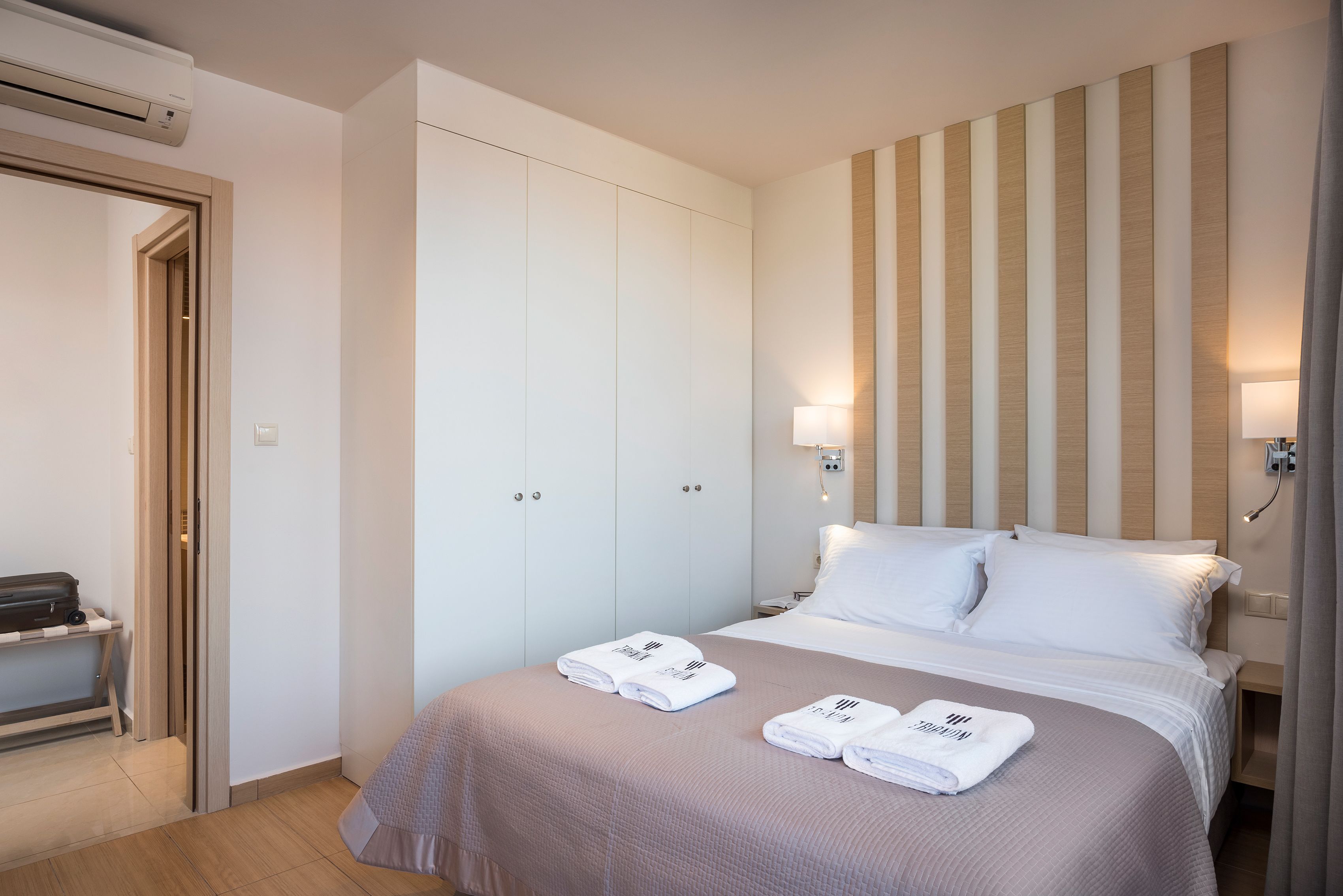 A hotel room with a bed, two wardrobes, a bedside table, a lamp, and a suitcase on a bench.