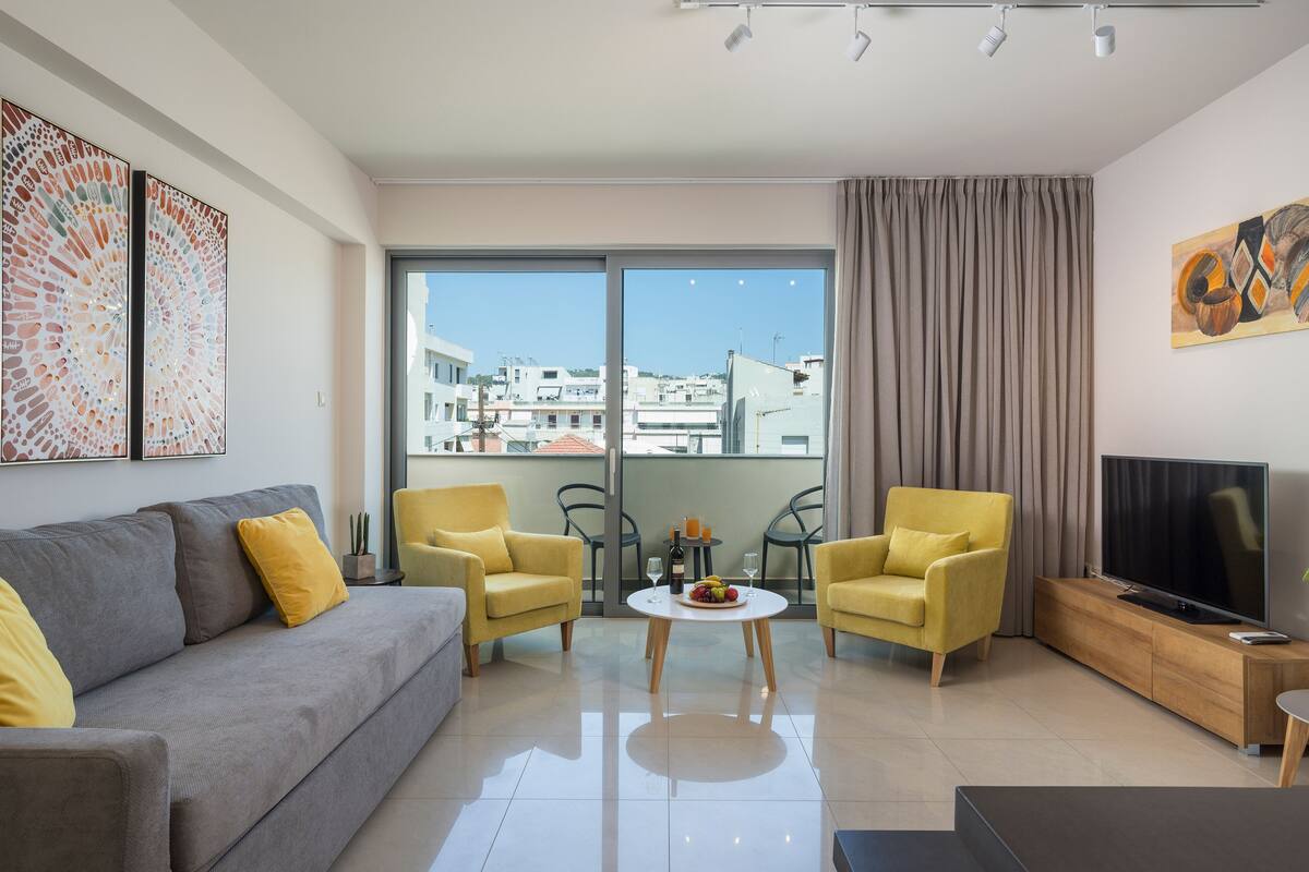 Una sala de estar moderna con un sofá gris, sillones amarillos, una mesa de centro redonda y un televisor de pantalla plana sobre un soporte de madera.