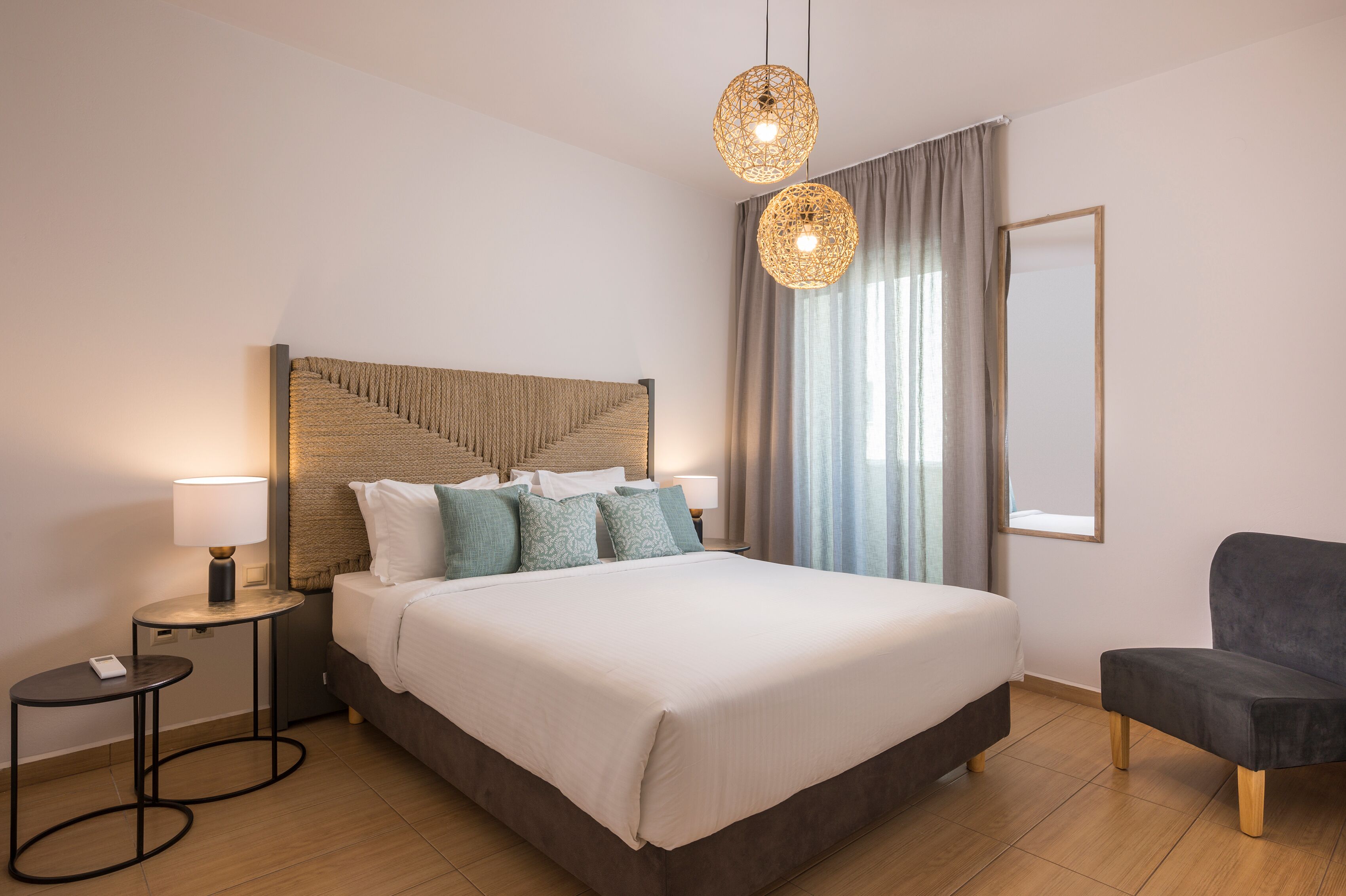 A modern hotel room with a large bed, a bedside table with a lamp, a chair, and a large window with curtains.