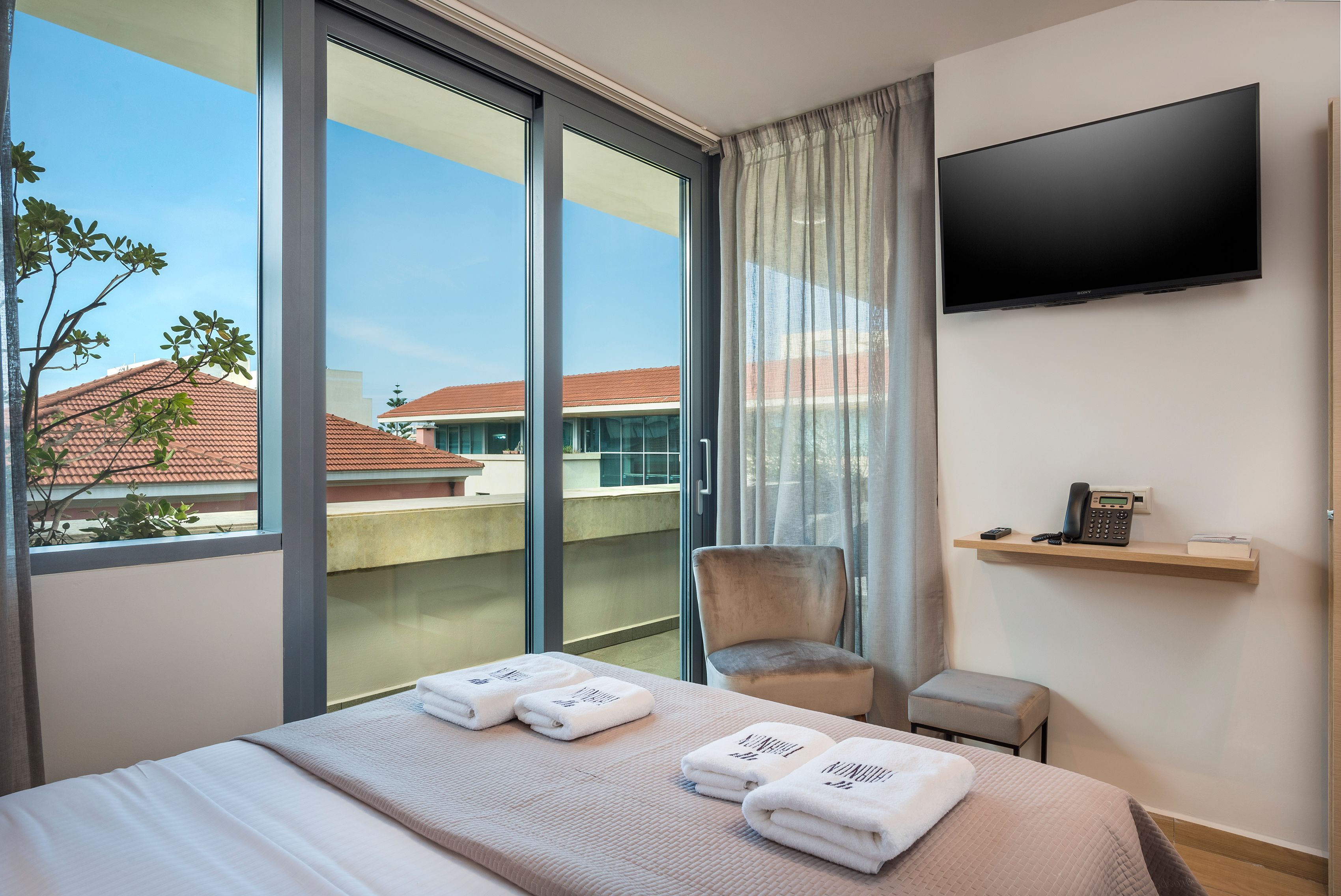 A hotel room with a bed, a chair, a TV, and a balcony with a view of buildings.