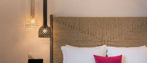 A neatly made bed with white and pink pillows, a laptop on a small wooden table beside it, and two woven wall hangings above the bed.