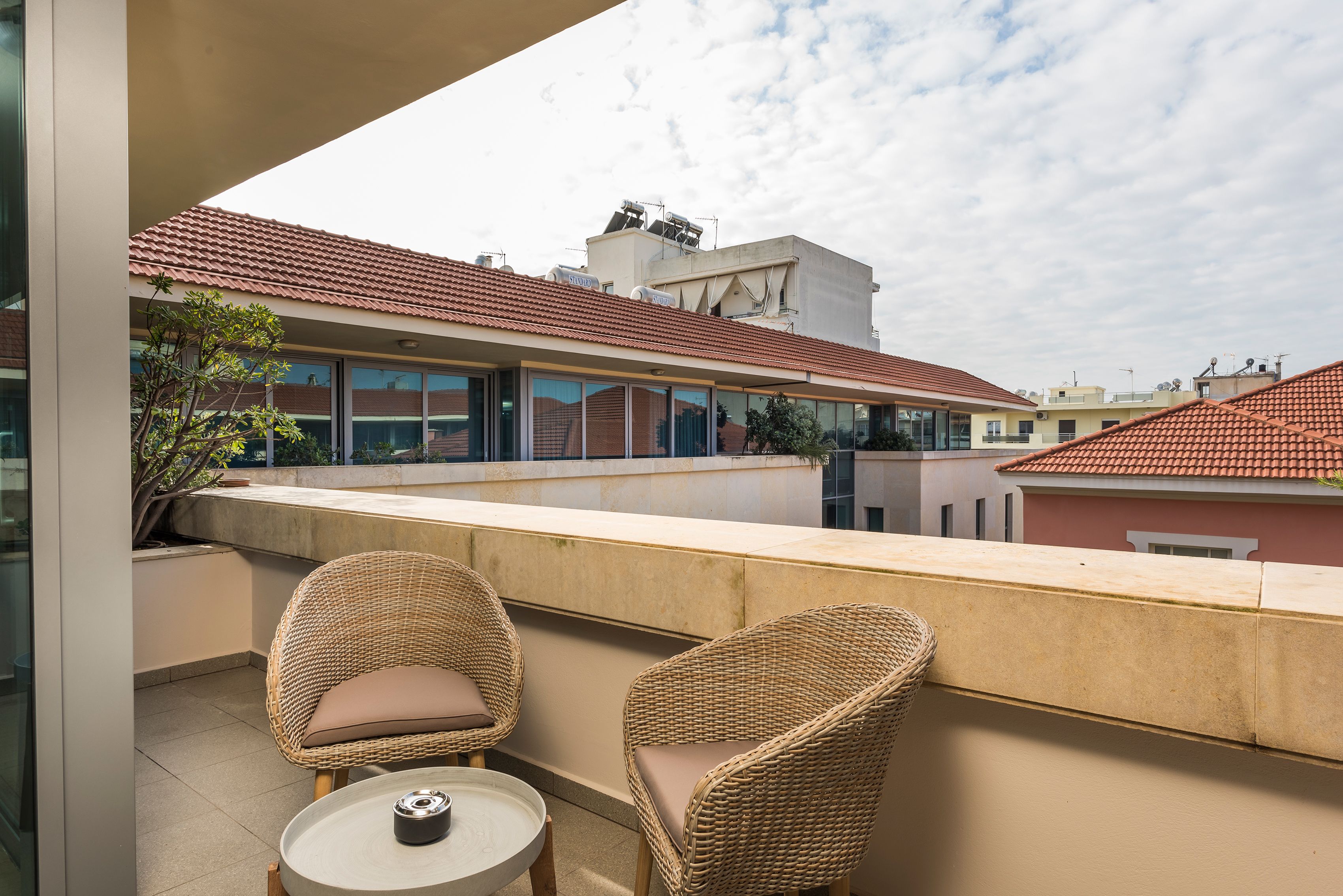 A balcony with wicker chairs and a small table, overlooking a residential area with red-tiled roofs and modern buildings.