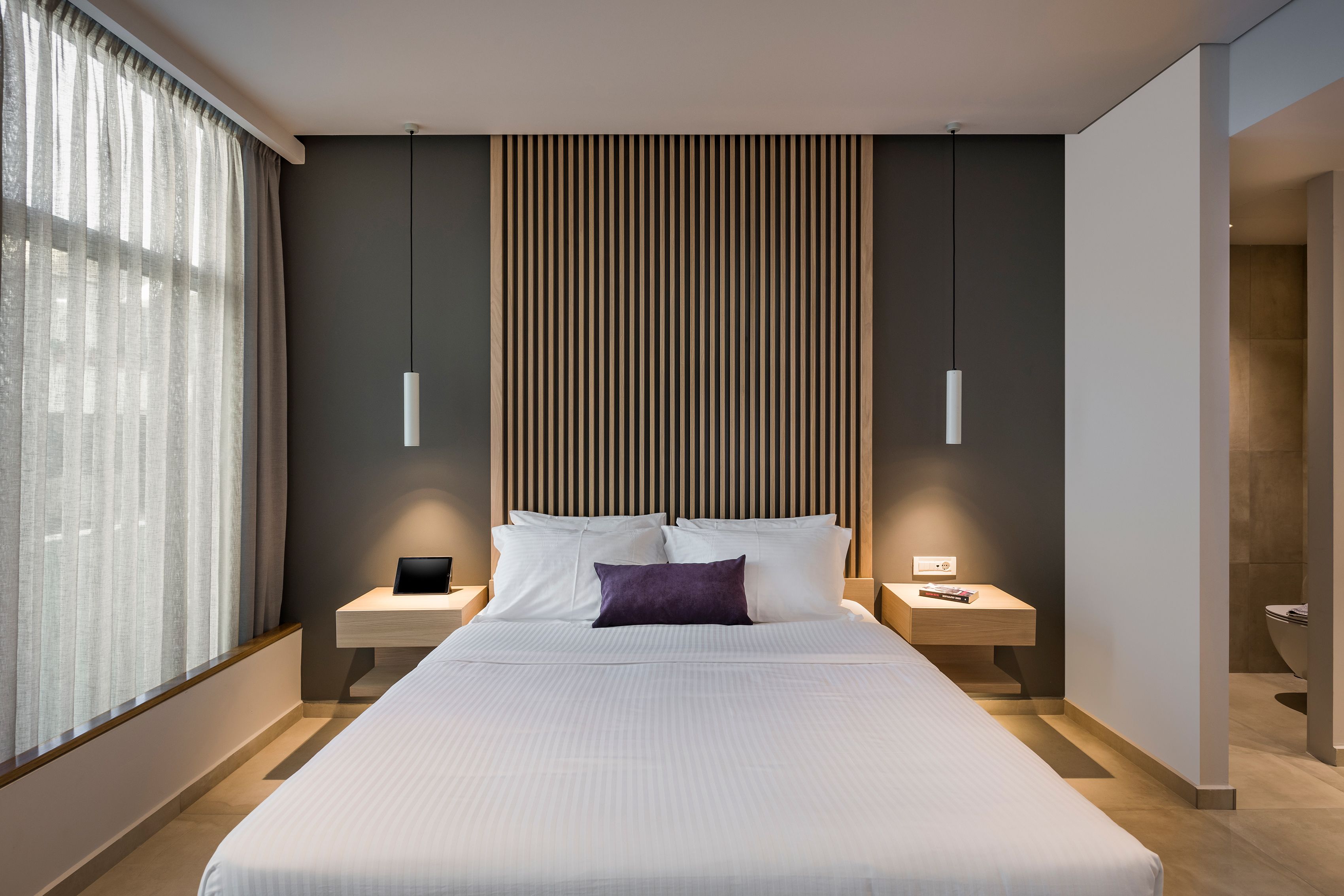A modern hotel room with a large bed, two bedside tables, a nightstand, a wooden slat headboard, and a large window with sheer curtains.