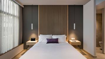A modern hotel room with a large bed, two bedside tables, a nightstand, a wooden slat headboard, and a large window with sheer curtains.