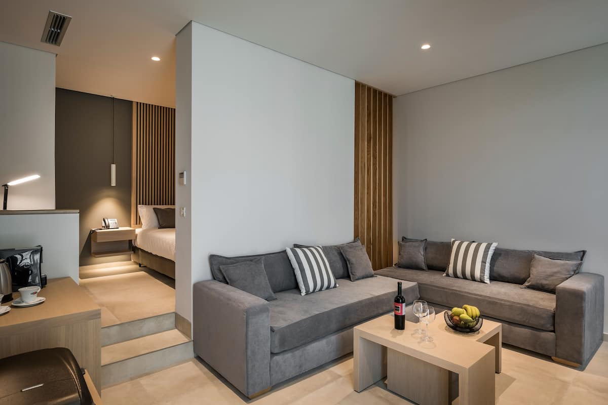 Una habitación de hotel moderna con un sofá gris, una pared de acento de madera, una pequeña mesa de comedor y una cama con mesita de noche.