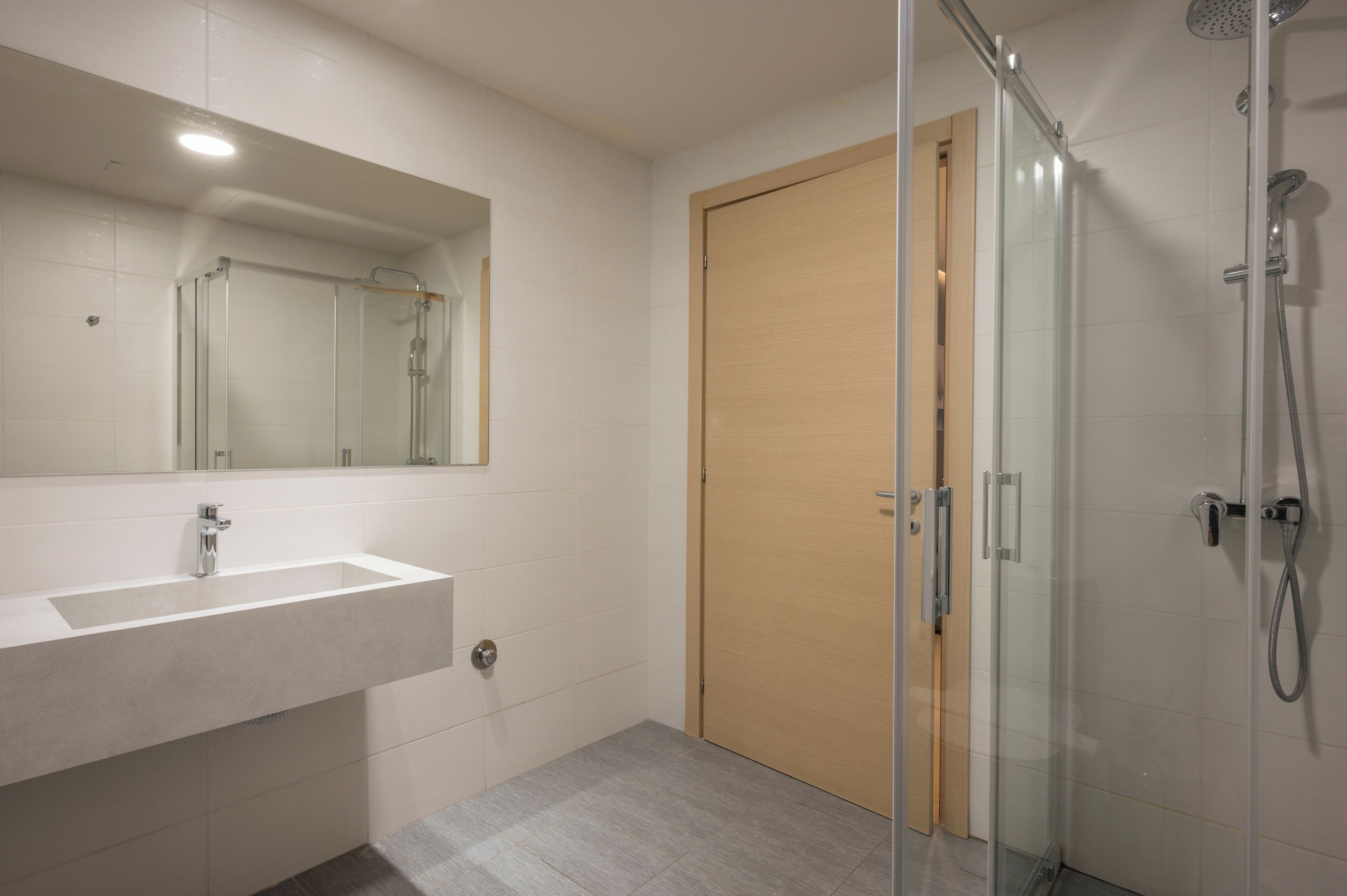 Une salle de bain moderne avec une douche à parois de verre, une porte en bois et un grand miroir au-dessus d’un lavabo blanc.
