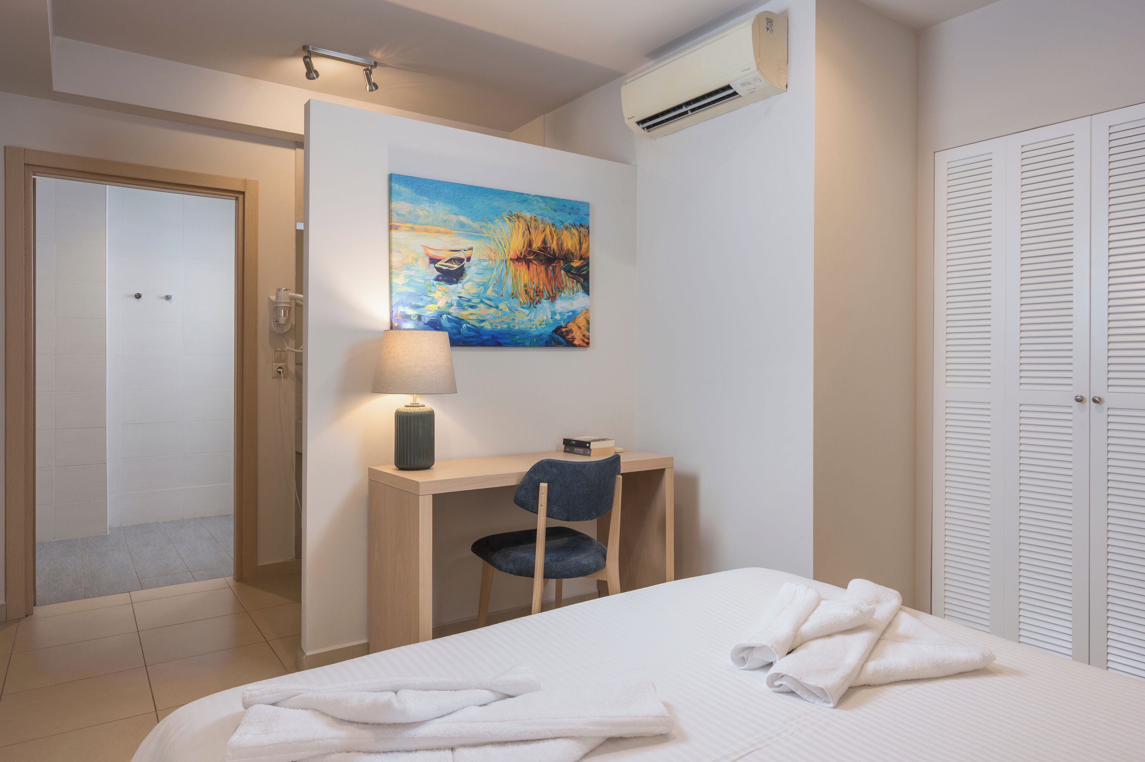 A hotel room with a bed, a desk with a chair, a painting on the wall, and a wardrobe.