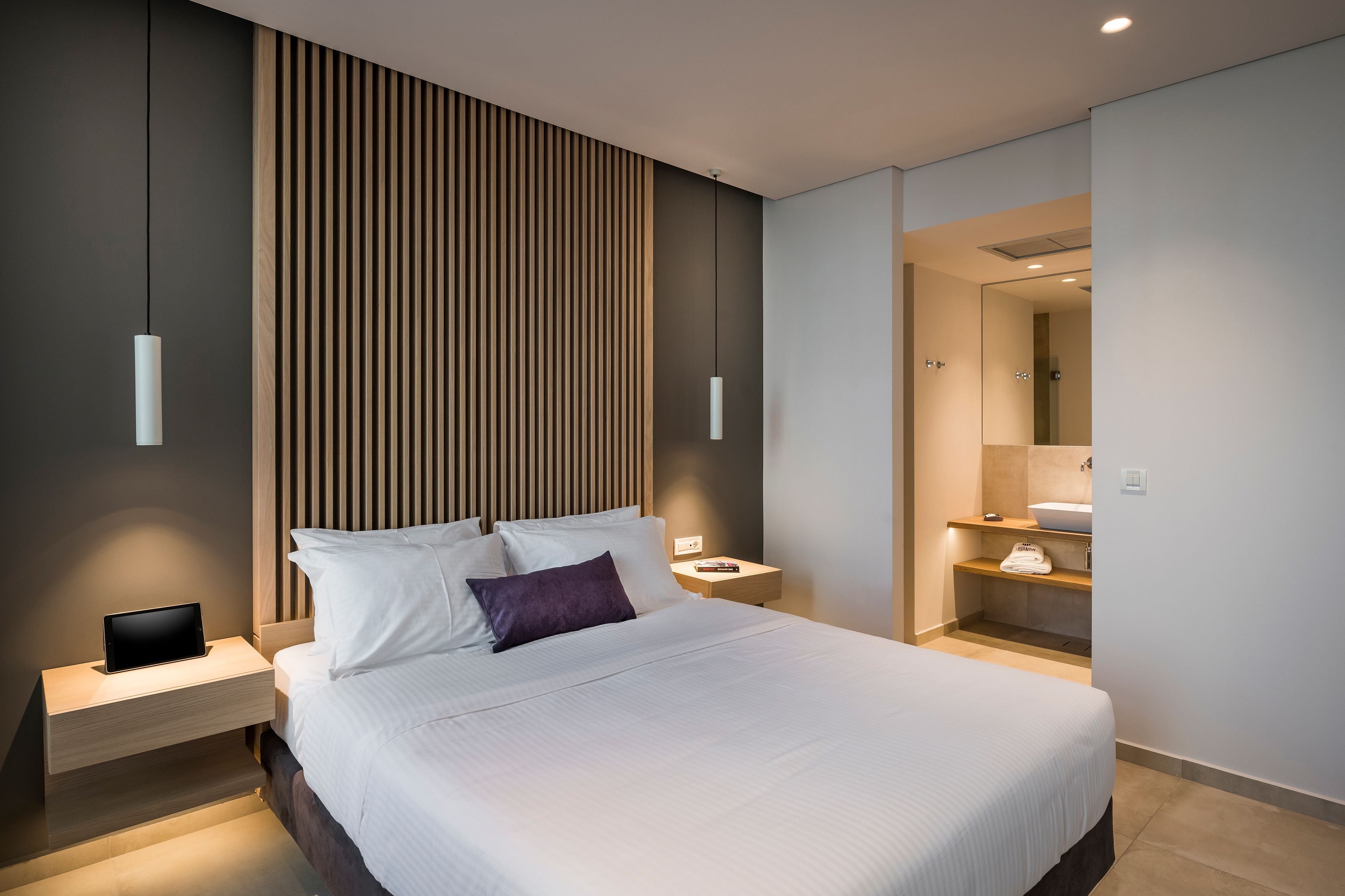 Une chambre d’hôtel moderne dotée d’un grand lit, de tables de chevet, d’une lampe de chevet et d’une étagère intégrée avec un miroir et un lavabo.