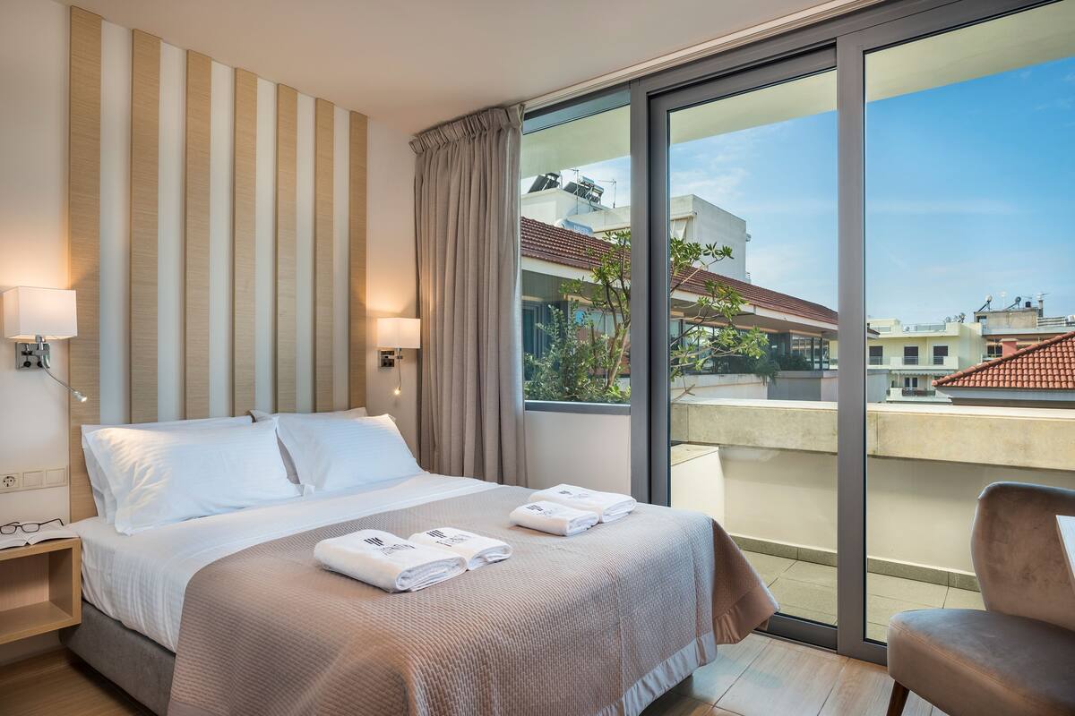 Habitación de hotel con una cama grande, dos mesitas de noche, una ventana con cortinas y vistas a edificios.
