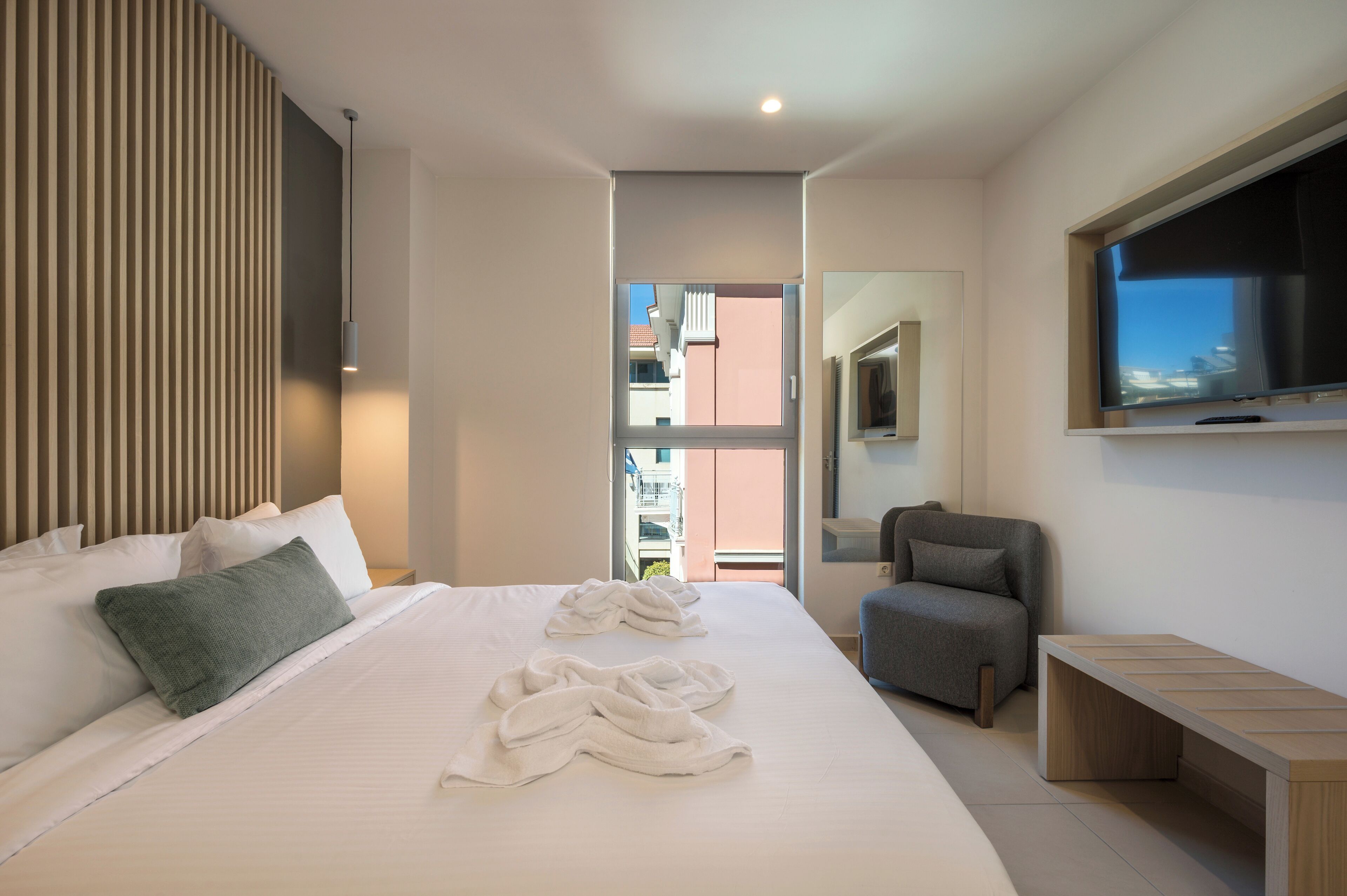 Een moderne hotelkamer met een groot bed, een grijze fauteuil, een aan de muur bevestigde televisie en een balkon met uitzicht op gebouwen.